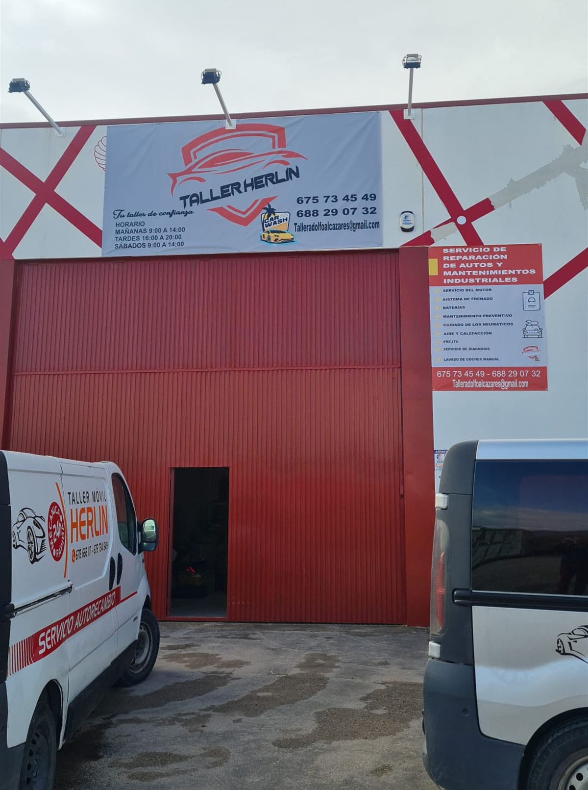 Fachada de Taller Herlin: nave con persiana roja, lona del rótulo y la furgoneta del taller móvil aparcada delante